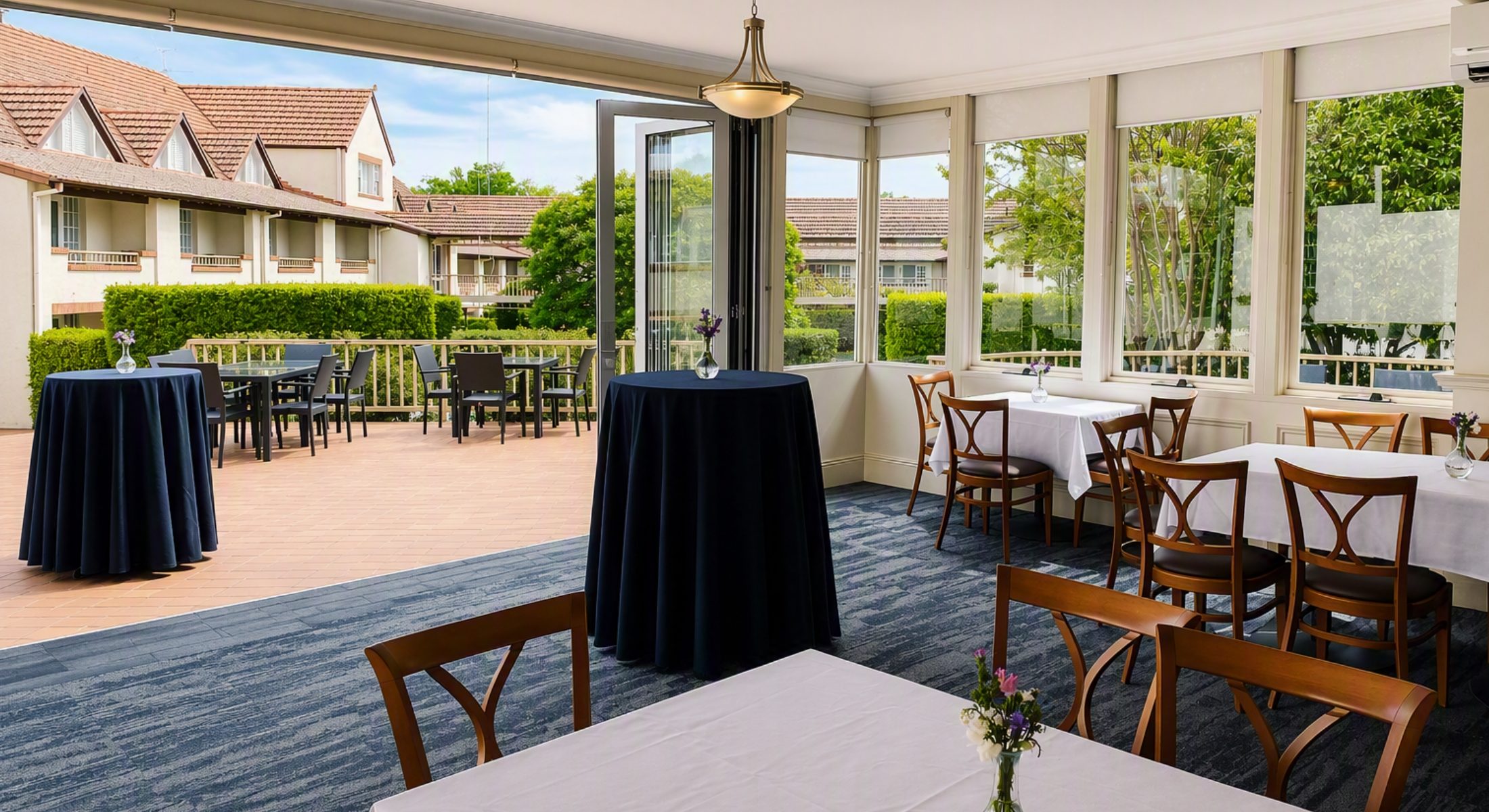 Courtyard Restaurant at Mercure Canberra featuring elegant dining tables with white linen, floor-to-ceiling windows overlooking courtyard gardens, and a light-filled function space opening onto an outdoor terrace.